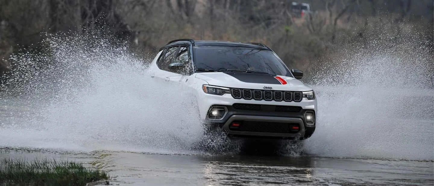 2026 Jeep Compass splashing through shallow water on a rugged trail with spray rising around the front of the vehicle.